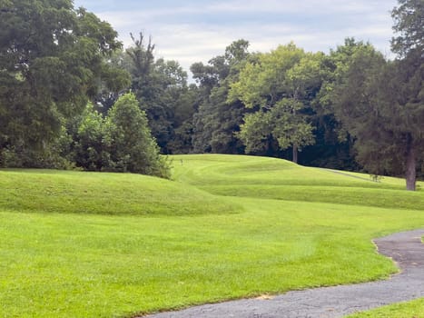 Walking path leads past curving body of a prehistoric Native American snake earthwork effigy mound at Great Serpent Mound in Ohio. Woodland background under a dramatic sky. Green grass is neatly trimmed and covers the curves of the snake. No people.