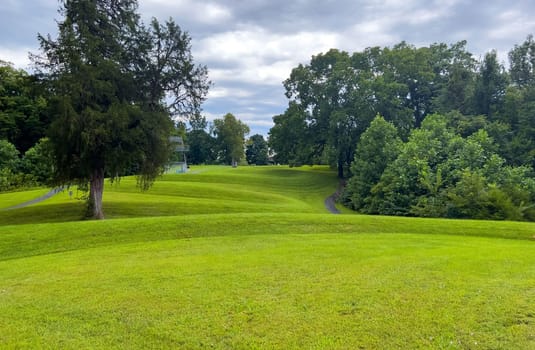 Curving body of a prehistoric Native American snake earthwork effigy mound at Great Serpent Mound in Ohio. Observation tower in background under a dramatic sky. Green grass is neatly trimmed and covers the curves of the snake. No people.