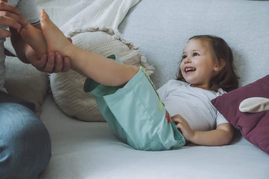 Father plays with little daughter 2-4 on couch. Dad tickles kids feet. Family having fun