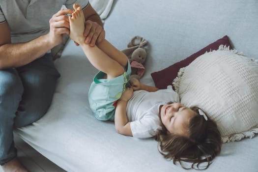 Father plays with little daughter 2-4 on couch. Dad tickles kids feet. Family having fun