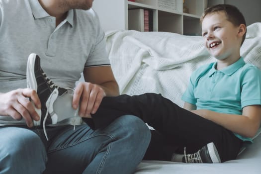 Dad and son sitting on couch. Father helps child to take off or put on shoes, kid laughs