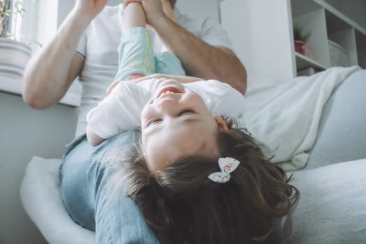 Dad and little girl 2-4 play on the couch. Kid lies on father's lap with her head down and laughs