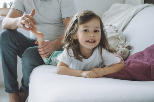 Father plays with little daughter 2-4 on couch. Dad tickles kids feet. Family having fun