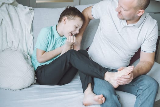 Father plays with his son 7-10 sitting on couch. Dad tickles kids feet. Family having fun