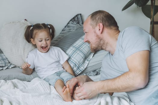Father plays with little daughter 2-4 on bed. Dad tickles kids feet. Family having fun