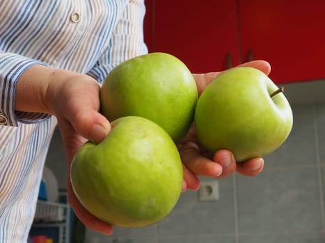 Apples in hand. The woman holds three green apples in her hand and offers them to the viewer. Organic food. Fresh fruits for food. Big sour apple. White shirt with stripes. Kitchen with red furniture.
