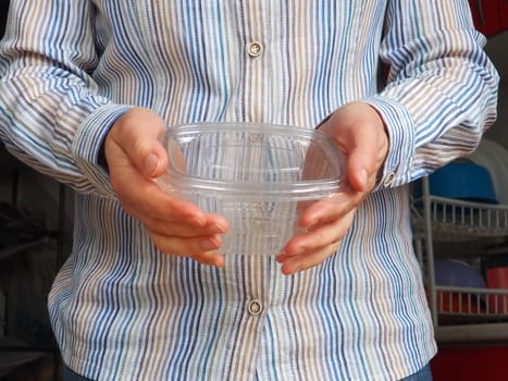 Female hands hold a plastic container or packaging. Kitchen utensils. A woman in a white striped shirt stands in the kitchen and shows a disposable plastic box with a lid. Hygiene and recycling theme.