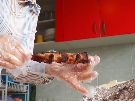 Grilled meat in disposable packaging and aluminum foil from the restaurant. Home delivery from restaurants and cafes. Female hands in disposable gloves hold meat on a stick