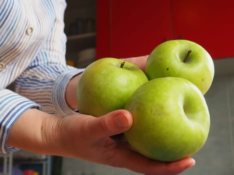 Apples in hand. The woman holds three green apples in her hand and offers them to the viewer. Organic food. Fresh fruits for food. Big sour apple. White shirt with stripes. Kitchen with red furniture.