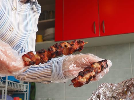 The grilled meat was delivered from the restaurant. Home delivery from restaurants. Women's hands in disposable gloves hold meat on a stick. Kitchen with red furniture. Cleanliness and safety of food.