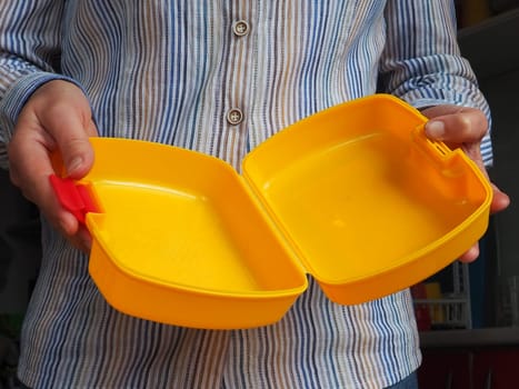 Female hands hold a yellow open plastic school lunch container or packaging. Kitchen utensils. A woman in a white striped shirt shows the lunch box. Food hygiene and recycling theme.