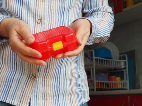Female hands hold a red closed plastic school lunch container or packaging. Kitchen utensils. A woman in a white striped shirt shows the lunch box. Food hygiene and recycling theme.