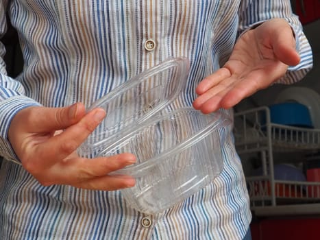 Female hands hold a plastic container or packaging. Kitchen utensils. A woman in a white striped shirt stands in the kitchen and shows a disposable plastic box with a lid. Hygiene and recycling theme.