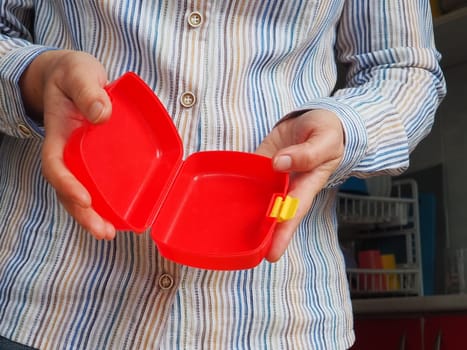 Female hands hold a red open plastic school lunch container or packaging. Kitchen utensils. A woman in a white striped shirt shows the lunch box. Food hygiene and recycling theme.