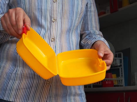 Female hands hold a yellow open plastic school lunch container or packaging. Kitchen utensils. A woman in a white striped shirt shows the lunch box. Food hygiene and recycling theme.