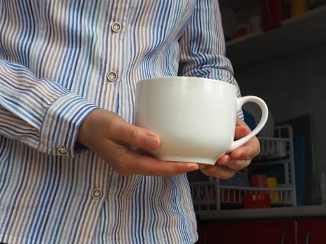 Female hands hold a large white ceramic mug or cup. Kitchen and cooking utensils, dishes. A woman in a white striped shirt stands in the kitchen and shows an unmarked cup. Copy space.
