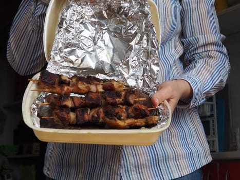 Grilled meat in disposable packaging and aluminum foil from the restaurant. Home delivery from restaurants and cafes. Female hands in disposable gloves hold meat on a stick