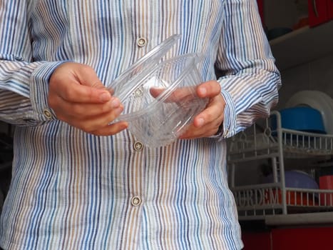 Female hands hold a plastic container or packaging. Kitchen utensils. A woman in a white striped shirt stands in the kitchen and shows a disposable plastic box with a lid. Hygiene and recycling theme.