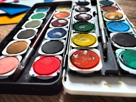 A set of watercolors of different colors for drawing. Multicolored round containers with bright paint. Tools for the lesson of drawing and labor. Colored paper. Red, blue, yellow, green colors