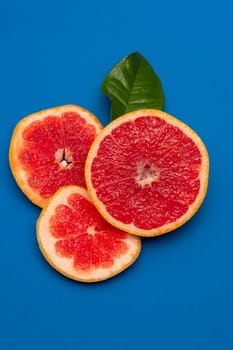 Sliced sides of a grapefruit, three pieces with a green leaf, red pulp isolated on a blue background, vertical shot