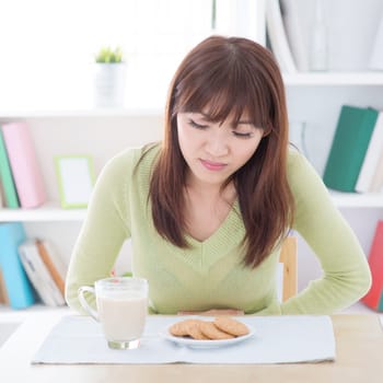 Woman holding stomach and suffering stomachache, while having dairy milk and cookies as breakfast, sitting at dining table.Young people indoors living lifestyle at home.