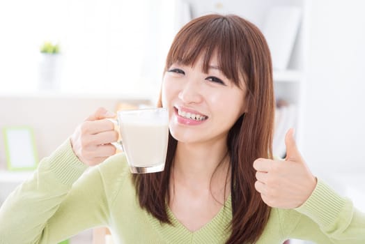 Portrait of happy Asian girl thumb up while drinking milk as breakfast. Young woman indoors living lifestyle at home.