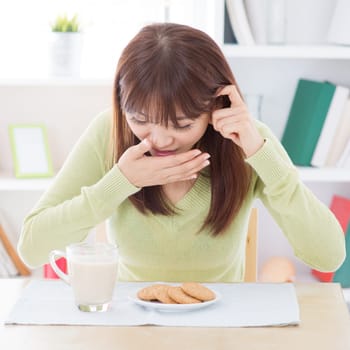 Asian woman feeling sick while having breakfast, milk and cookies on dining table. Young girl indoors living lifestyle at home.