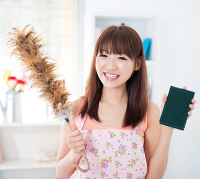 Happy Asian housewife with apron housecleaning, hand holding duster and cleaning sponge. Young woman indoors living lifestyle at home.