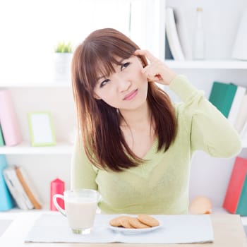Portrait of Asian girl thinking while having breakfast, milk and cookies on dining table. Young woman indoors living lifestyle at home.