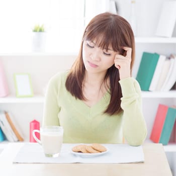 Asian woman feeling bored with her everyday breakfast, milk and cookies on dining table. Young people indoors living lifestyle at home.