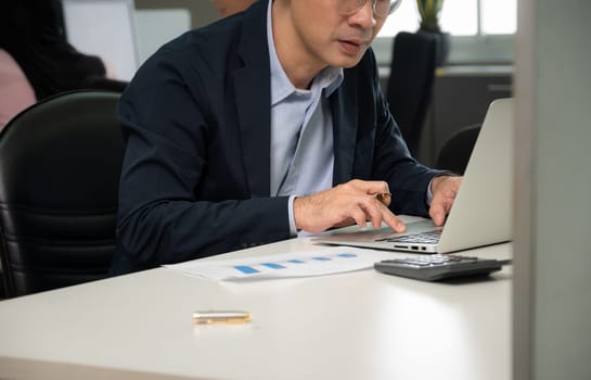 Young Asian businessman in eyeglasses working on laptop at his desk in open space office with colleagues in the background