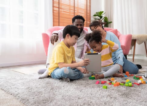 Happy black little curly girl with her family looking at tablet and making online video call at home. Four people sitting on rug floor carpet in living room interior.