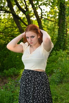 A pretty young red-haired woman, wearing a light-colored blouse, arms on her head, in green nature
