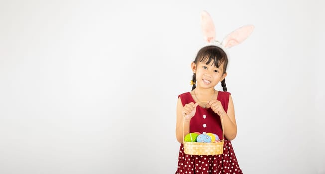 Happy Easter Day. Smile Asian little girl wearing easter bunny ears holding basket of full colorful eggs smiles broadly isolated on white background with copy space, Happy child in holiday