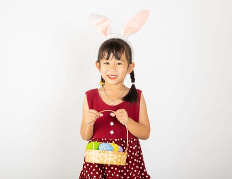 Happy Easter Day. Smile Asian little girl wearing easter bunny ears holding basket of full colorful eggs smiles broadly isolated on white background with copy space, Happy child in holiday