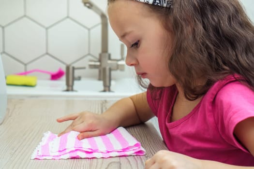 Pretty little girl wipe kitchen table with household rag for cleaning. Portrait closeup of child tidying up kitchen side view. Home wet cleaning concept. Kid help clean house. Room cleaning supplies.