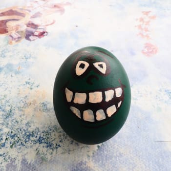 Easter egg dyed green with a painted laughing face. Cool grimace with eyes and big white teeth. Scary mug for Halloween. Emoticon for Easter. Light abstract background