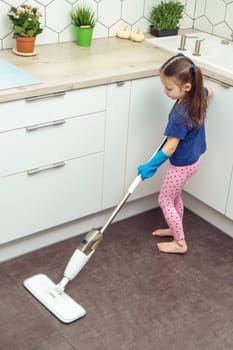 Nice little girl in household gloves wash floor with modern mop with removable brush near kitchen set. Full length photo of child tidying up kitchen from above view. Home wet cleaning concept.