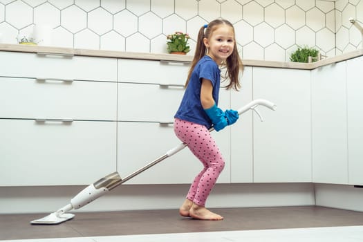 Happy little girl sitting on modern mop for floor washing with removable brush near kitchen set. Full length photo of child tidying up kitchen side view. Home cleaning concept. Kid help clean house.