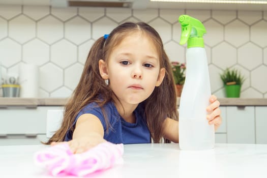 Pretty little girl with detergent sprayer and household rag wipe table. Portrait of child tidying up kitchen, home cleaning concept. Kid help clean up house. Room cleaning supplies, cleanser.