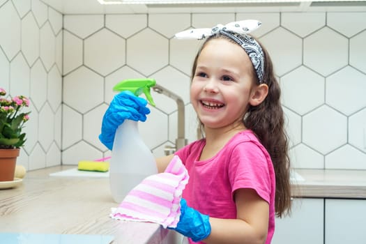 Joyful little girl in protective gloves wipe kitchen set with household rag and detergent sprayer. Portrait of happy child tidying up kitchen. Home wet cleaning. Detergent and cleaning supplies.