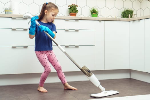 Pretty little girl in household gloves wash floor with modern mop with removable brush near kitchen set. Full length photo of child tidying up kitchen side view. Home wet cleaning concept.