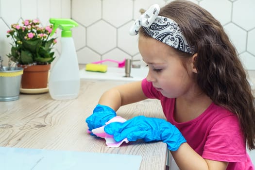 Pretty little girl in protective gloves wipe kitchen table with household rag for cleaning. Portrait of child tidying up kitchen side view. Home wet cleaning concept. Detergent and cleaning supplies.