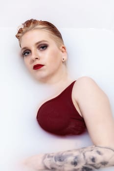 Attractive young woman with blue eyes, red lips and tattoo is lying in spa bath with milk and looking at camera.  Vertically.