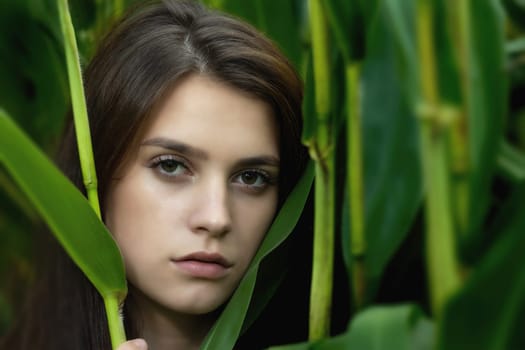 Beautiful tanned young woman is posing among the green plants. Horizontally.