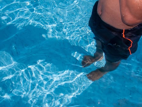 High angle view of torso and legs of a teenage boy standing in clear blue swimming pool water wearing swimwear. Shot in natural summer sunlight, his shadow is on the pool floor with waves and ripples. With copy space.