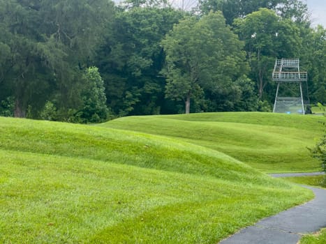Walking path leads past curving body of a prehistoric Native American snake earthwork effigy mound at Great Serpent Mound in Ohio. Observation tower in background under a dramatic sky. Green grass is neatly trimmed and covers the curves of the snake. No people.