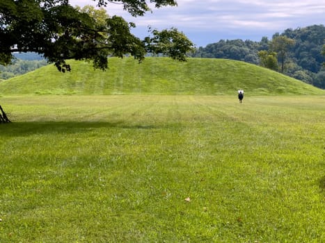 Grass covered prehistoric native American Hopewell culture burial mound at Siep Earthworks in Ohio under a dramatic cloudy sky with green grass foreground. A person walks toward the sign at the base of the mound.