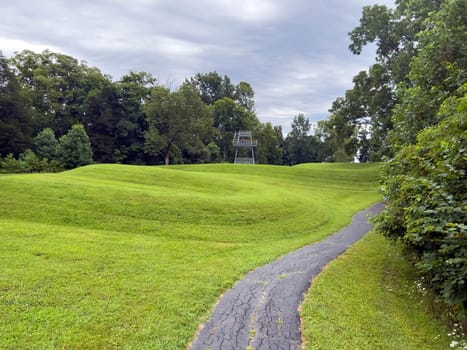 Walking path leads past curving body of a prehistoric Native American snake earthwork effigy mound at Great Serpent Mound in Ohio. Observation tower in background under a dramatic sky. Green grass is neatly trimmed and covers the curves of the snake. No people.
