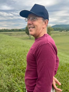 Caucasian senior man with glasses and a hat turns smiling toward camera on the top of a green grass covered hill under a dramatic sky. Active lifestyle and wellbeing concepts, as well as success and achievement.